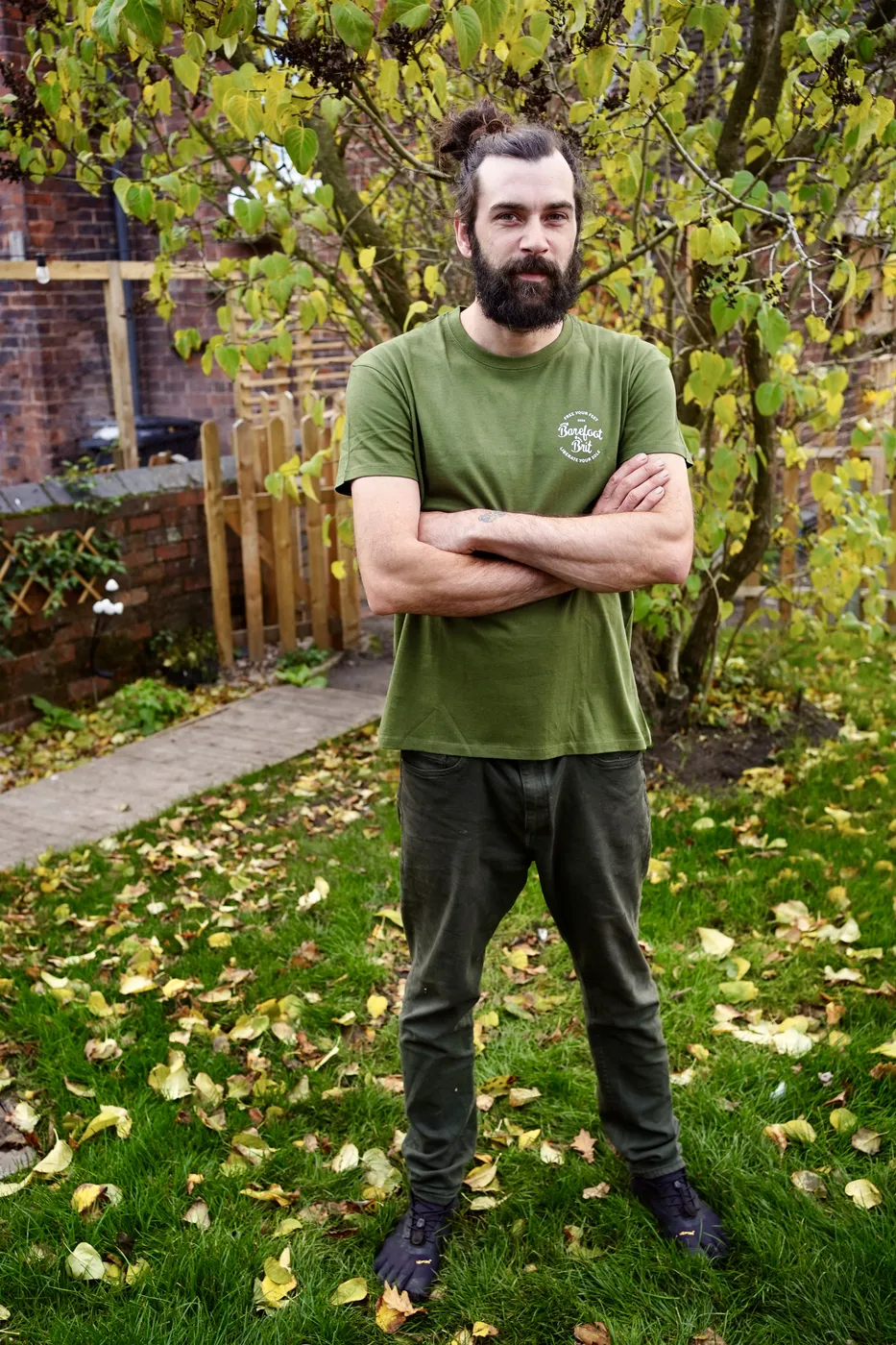 Dan standing in the garden in a green Barefoot Brit t-shirt and toe shoes, arms folded